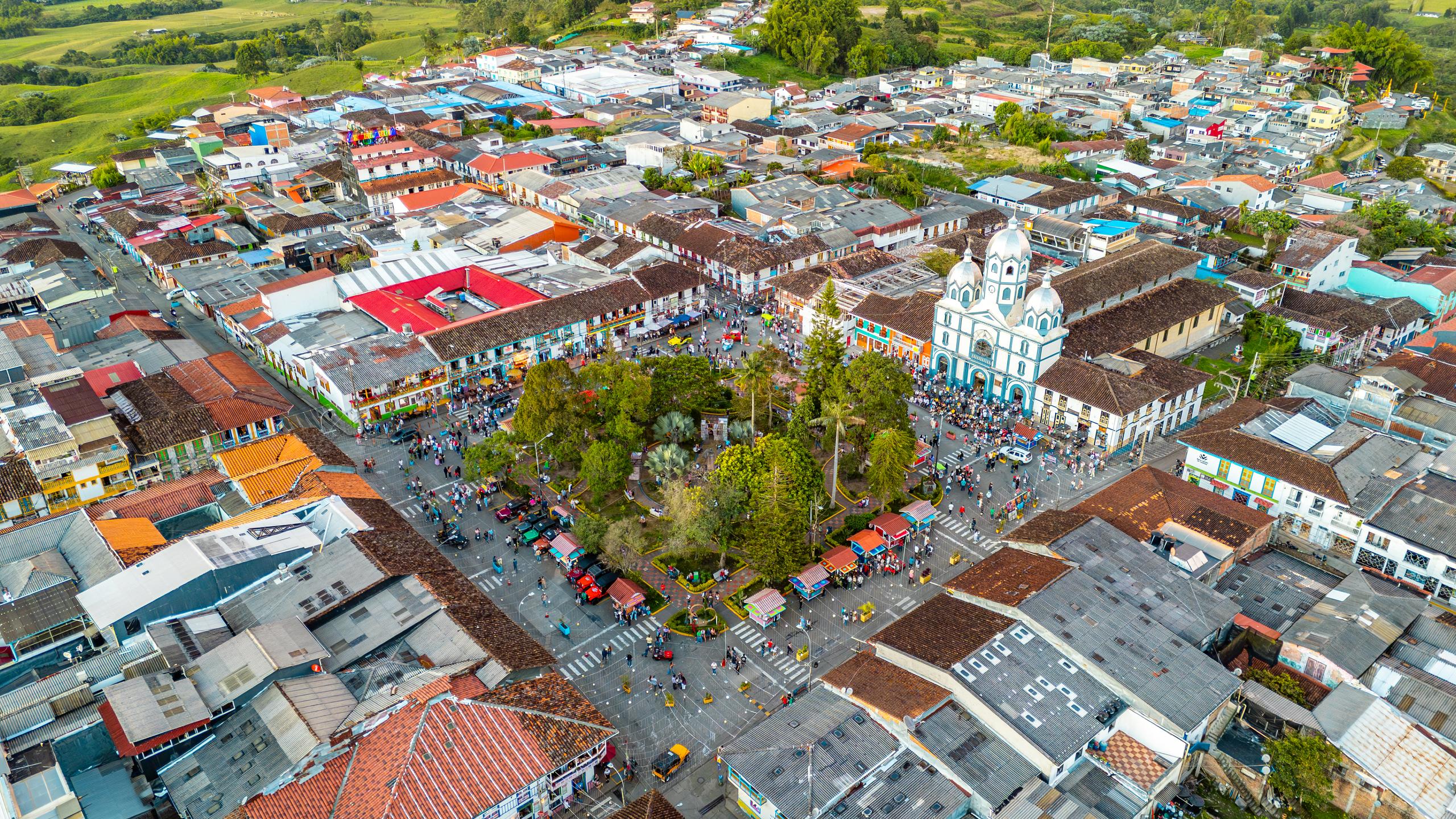 Vista aérea de la plaza principal de un pueblo del Eje Cafetero colombiano: parque central arbolado, iglesia blanca y azul, tejados de colores y montañas verdes al fondo.